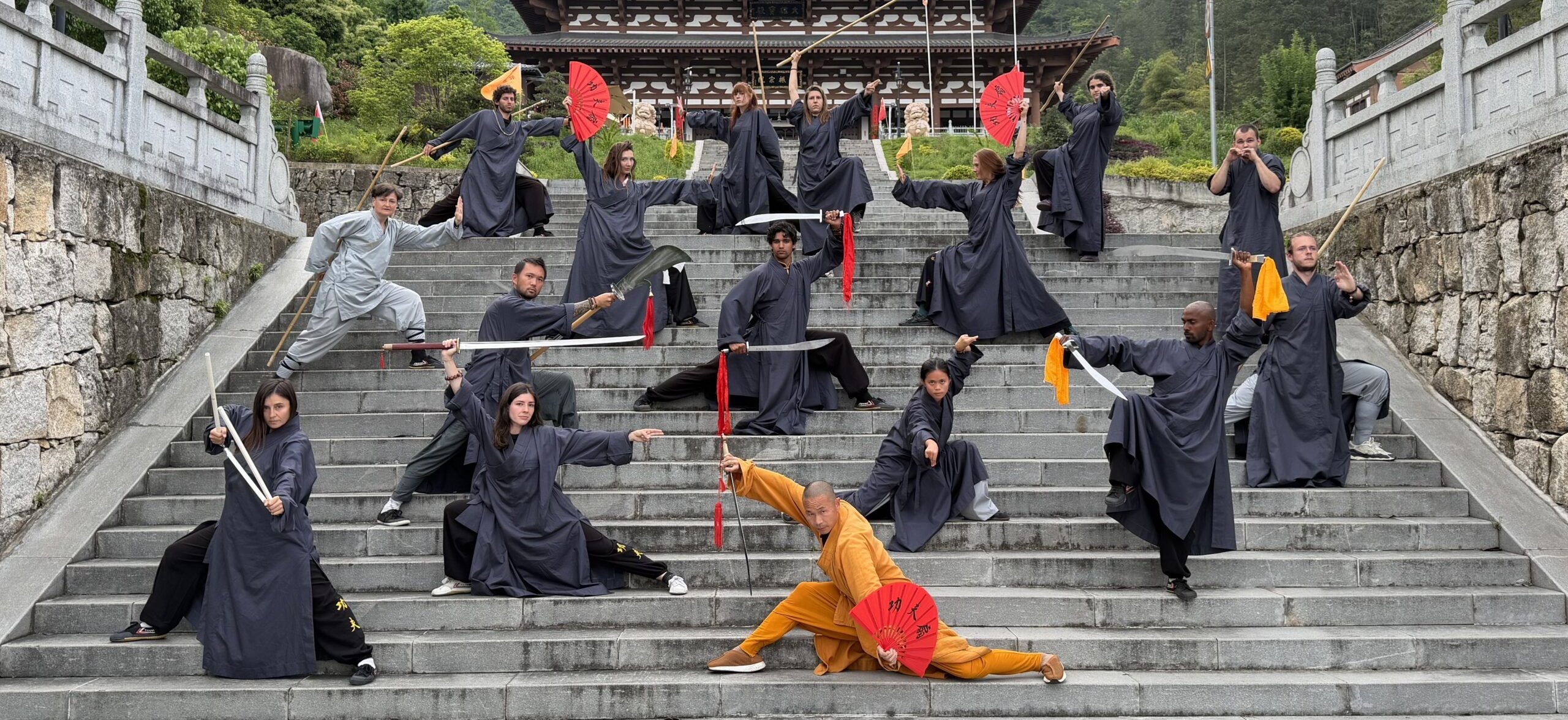 The cohort posing with swords, spears, and red silk fans on the temple steps, Shifu in a gold robe at the center, mountains behind